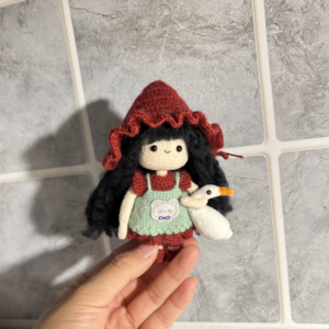 A handmade crochet goose girl doll with braided hair, standing on a wooden base and gently holding a small crochet goose in front of her.