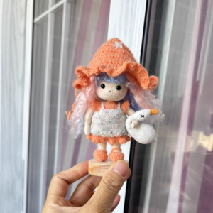 A handmade crochet goose girl doll in traditional dress, standing on a small wooden base and cradling a tiny crochet goose in her arms.