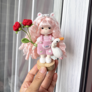 A handmade crochet goose girl doll standing on a wooden base, dressed in traditional clothing with braided hair and holding a small goose.