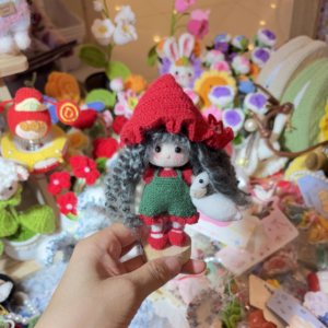 A close-up photo of a handmade crochet goose girl doll displayed on a small wooden base, showing her detailed braided hair, embroidered facial features, and the tiny goose she holds.