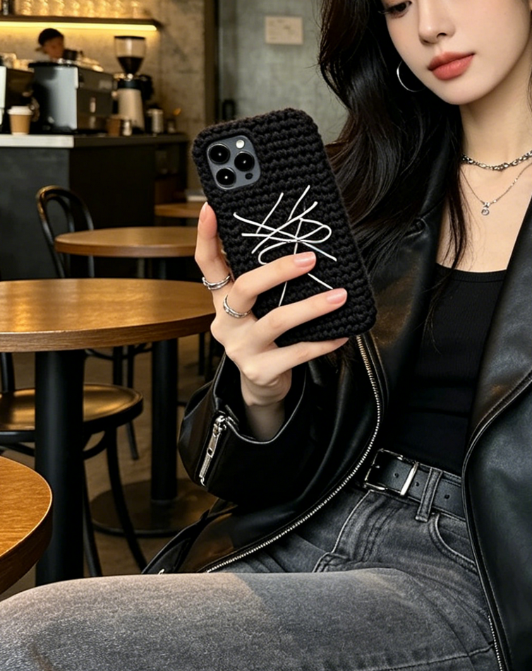Industrial cafe lifestyle shot, woman holding black hand crochet phone case with white sewn abstract bow, grunge streetwear handmade phone sleeve gift for her