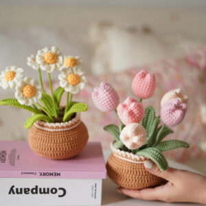 A complete flower potted crochet kit displayed on a white surface, including a small terracotta-colored pot, green yarn for leaves, colorful yarn for flowers, a wooden crochet hook, stuffing, and a finished sample flower.