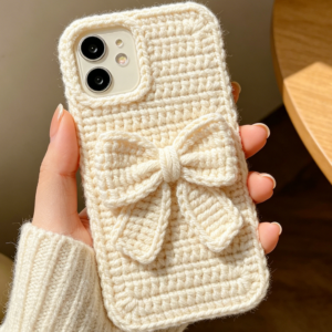 Handmade crochet phone case featuring a cute bow design in soft pastel pink, with textured stitch details and a secure fit around the smartphone, displayed on a light wooden surface.