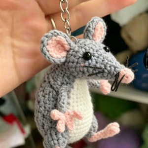 Handmade crochet mouse pendant in soft gray and pink, featuring round ears, a tiny pink nose, embroidered eyes, and a small tail, hanging from a delicate cord against a clean white background – perfect as a bag charm or keychain.